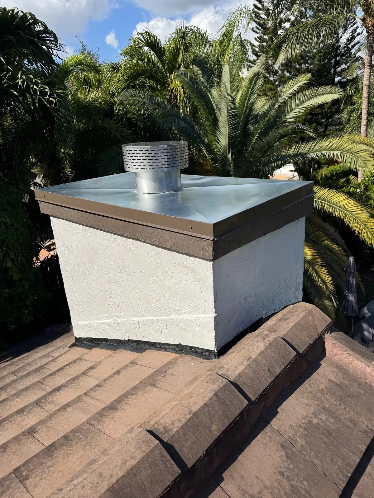 Finished white stucco chimney chase with a metal chase cover and a stainless steel cap on a tile roof, palm trees behind